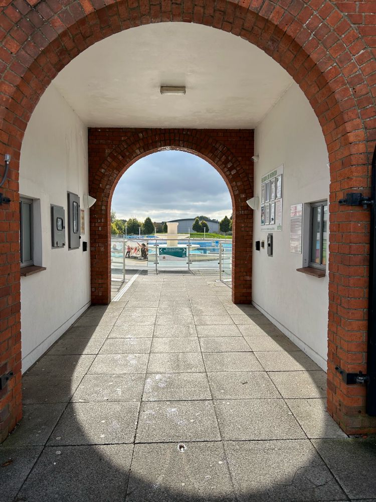 Entrance to Hillingdon leisure centre. Through an archway, we can see an inviting outdoor pool and large fountain beyond.