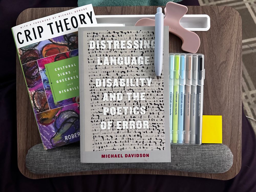 Overhead view of a brown lap table on a couch cushion. From left to right: Robert McRuer’s book “Crip Theory: Cultural Signs of Queerness and Disability,” Michael Davidson’s book “Distressing Language: Disability and the Poetics of Error,” a blue uniball one p pen clipped to the second book, and pink book claw clip above that, and pack of Staedtler highlighters to the right and behind the second book, and small golden yellow post-its to the right of that.
