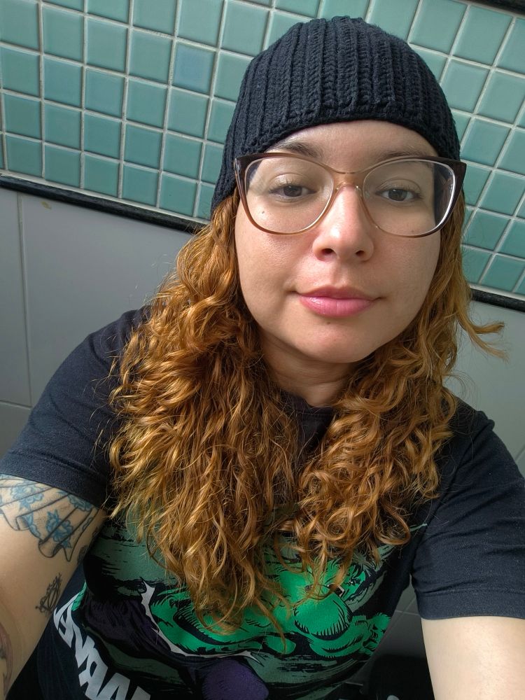 Foto minha: mulher branca, cabelo longo ruivo claro e ondulado emoldurando o rosto, uso óculos de grau em tom claro, blusa preta com um desenho do Hulk, tatuagens no braço esquerdo e usando uma touca de crochê na cor preta. Foto feita em um local fechado