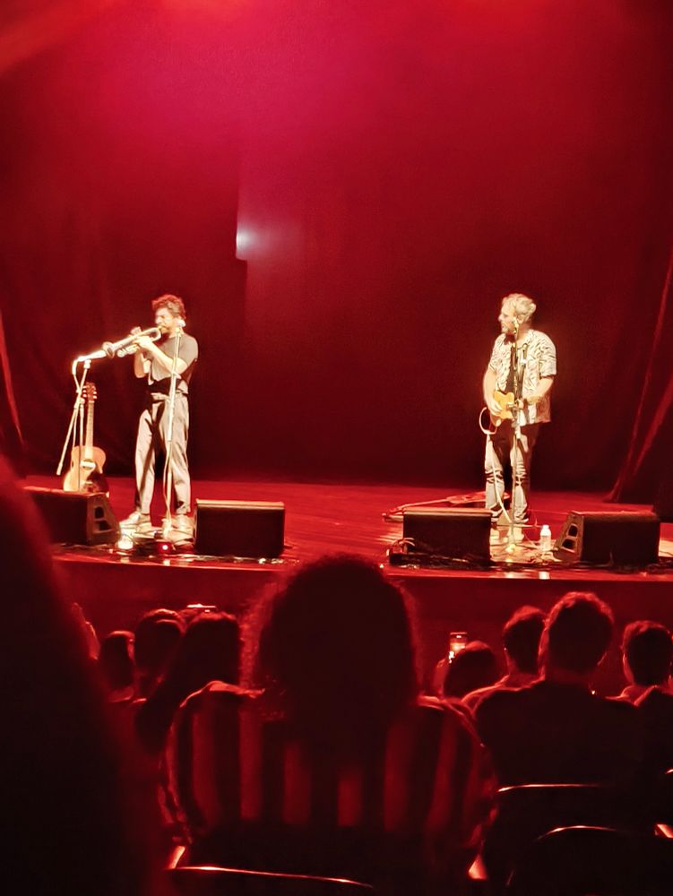 Imagem de um palco baixo, com dois artistas: um de cabelo preto altura mediana, camisa e calça preta, tocando um trompete. O outro um pouco mais alto, usando uma camisa branca estampada de botões abertas por cima de uma preta e calça preta, cabelo branco, tocando um violão. Microfones perto deles e luz vermelha no fundo. Pessoas de sentadas de costas olhando para eles na plateia de um teatro. 