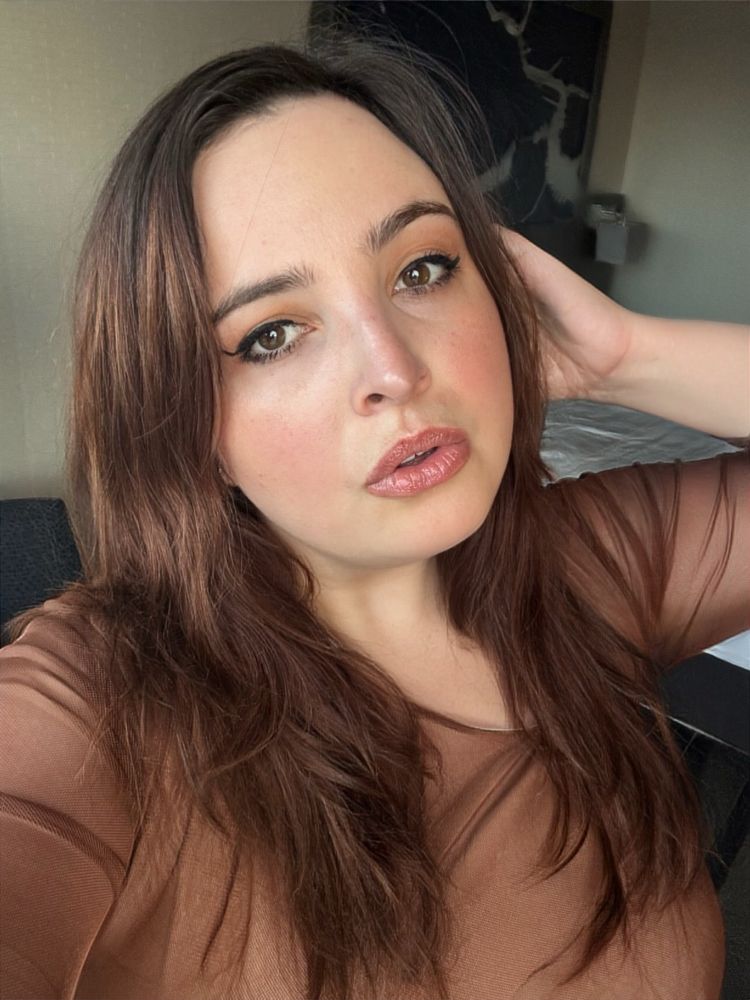Stella in soft natural light. Her cheeks aglow, wearing a brown mesh dress, her head tilted abd one hand placed on her auburn hair. 
