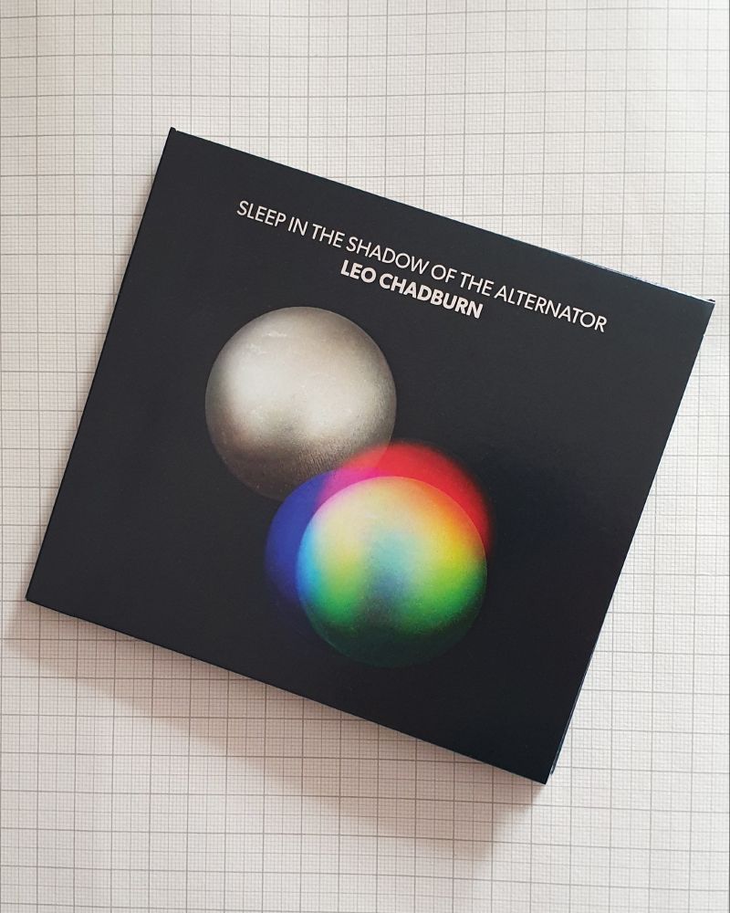 Cover of the CD version of Leo Chadburn, 'Sleep in the Shadow of the Alternator': two spheres of steel float in a black void, one glows red, green, blue.