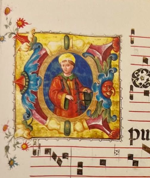 Pictorial detail from an illuminated manuscript page of C16th music, depicting a haloed, brown-haired, beardless male figure within a decorative letter C, against a blue background. He wears a red robe with green trim. His elbows are bent so that his forearms are in front of him, held perpendicular to his body. Toward the right side of the picture as the viewer sees it, his left hand rests on top of a gridiron, the symbol of his martyrdom. The letter C is contained within a square with a background of gold paint.