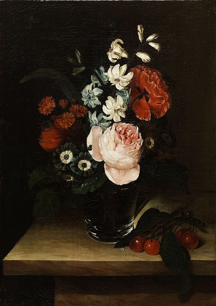A small bouquet in a clear glass or vase, sitting on a wooden table. The dominant flower colors are red and pink, with a little bit of white and blue. Next to the glass, to the viewer's right, sits a little branch of cherries. 