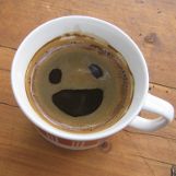 A top view of a coffee cup filled with coffee. The surface of the coffee features foam that resembles a smiling face, complete with two eyes and a wide mouth.
