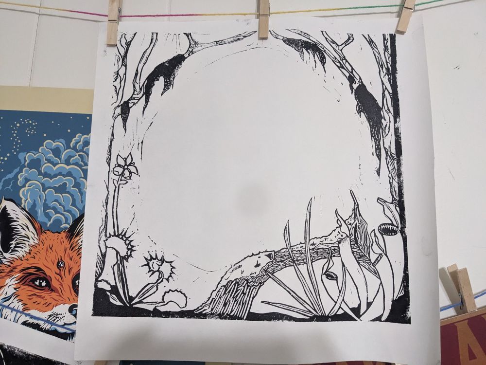 A drying print in black and white clothes pinned to yarn along a wall. The image is of a nature filled framing image. There is grass, pitcher plants, a mossy log, and Venus fly traps along the bottom. Around the sides are trees with Spanish moss dangling from the branches. 