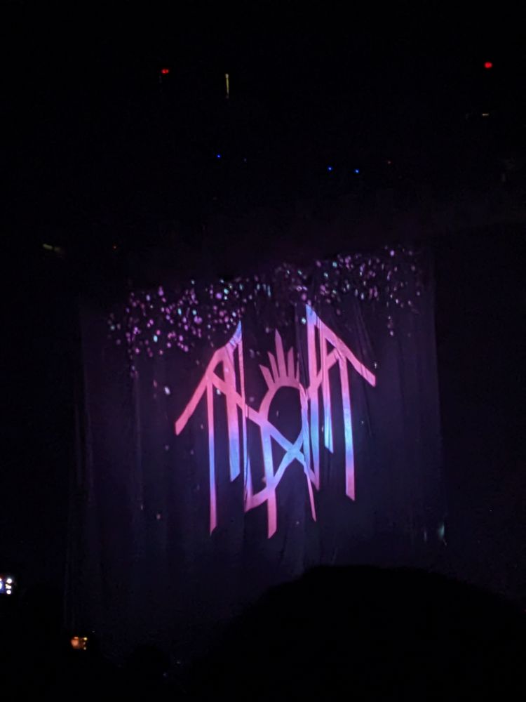 A close up of a stage with a curtain across it. The stage is dark and only the curtain is lit up. The lights project the Sleep Token logo in pinks and lavender.