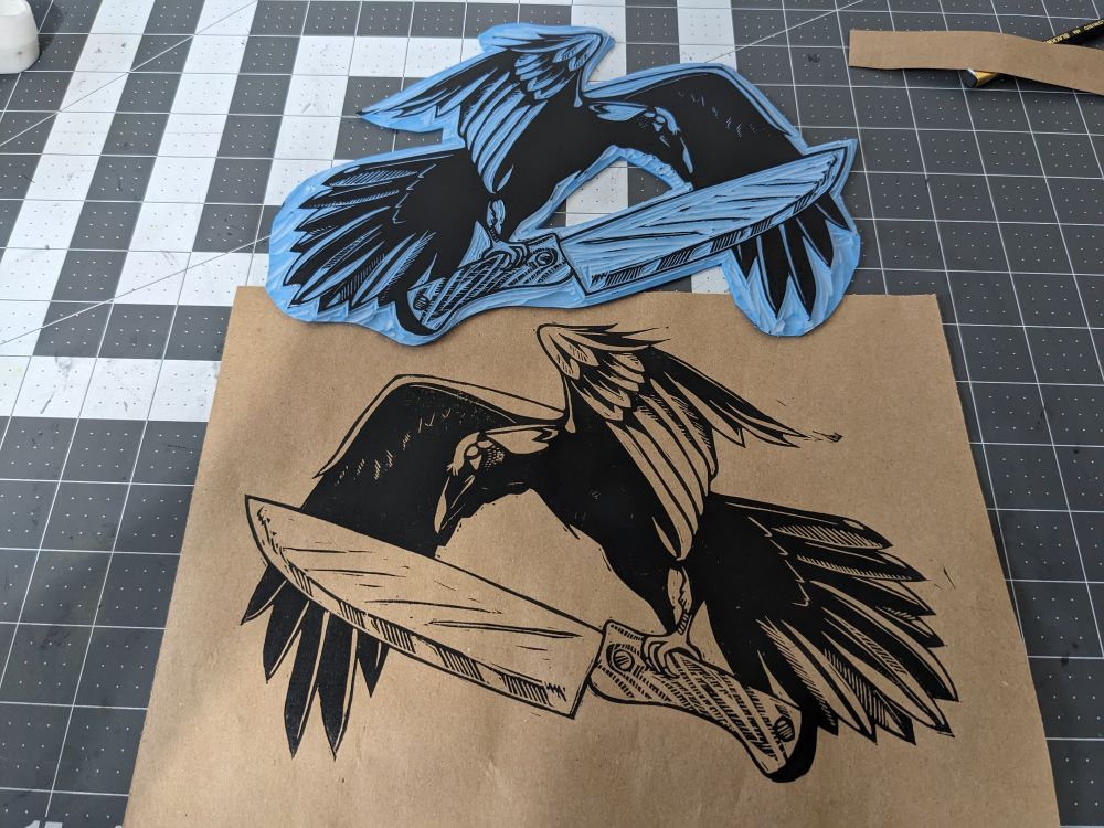 A block and the relief print made from it on a desk mat. The block is at the top, depicting a crow mid flight holding a wooden handled knife. Below it is the resulting print of the same design, reversed, printing in black ink on brown paper for the first artist proof print.
