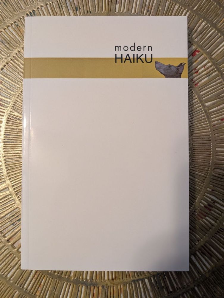 On a gold colored placemat, a white book rests in the center. In the first third of the book is a gold stripe with a doberman (I think) looking up at the title of the publication, "Modern Haiku"