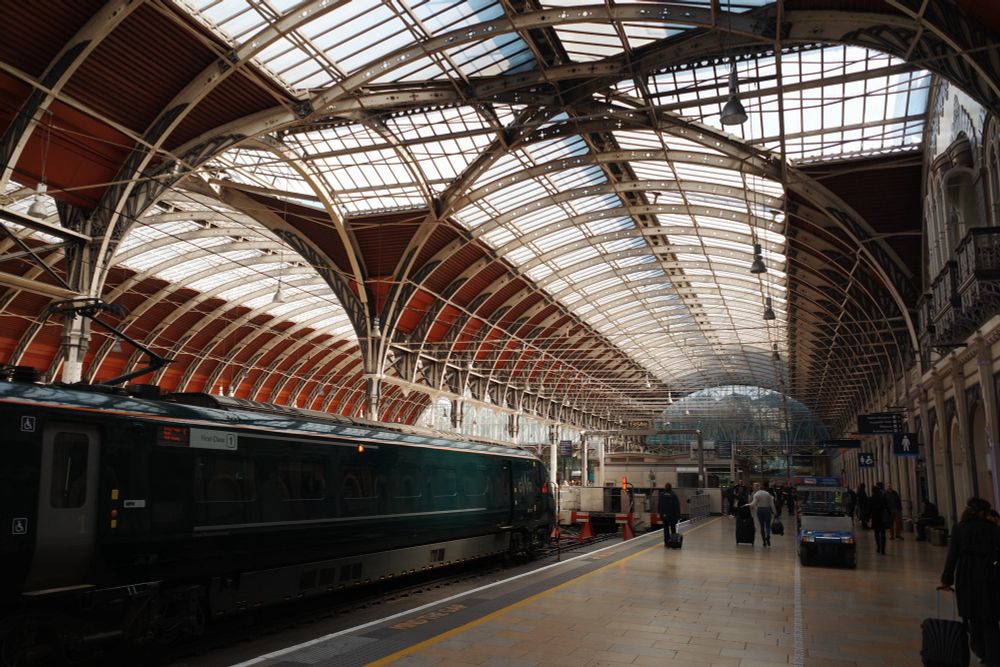 A train is stopped at a platform under the large arched glass-and-steel roof of Paddington Station in London. People walk along the platform, some pulling suitcases, beneath the natural light streaming in.
