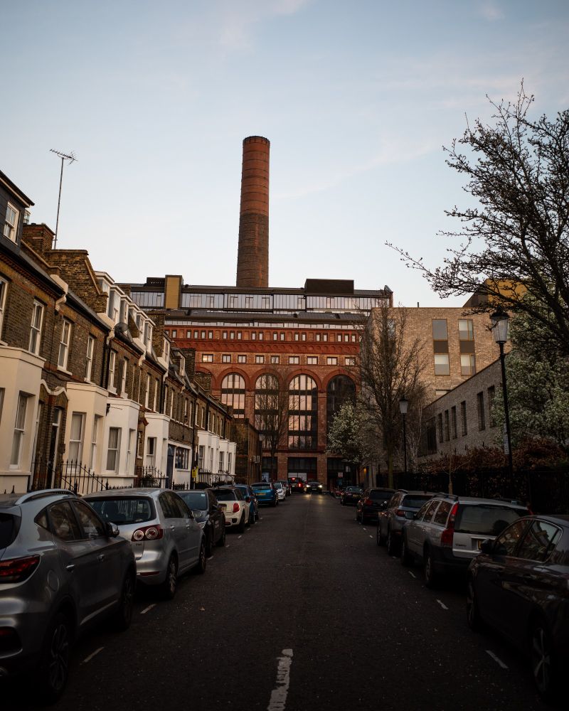 Residential street leading to a tall brick chimney under a pale blue sky.