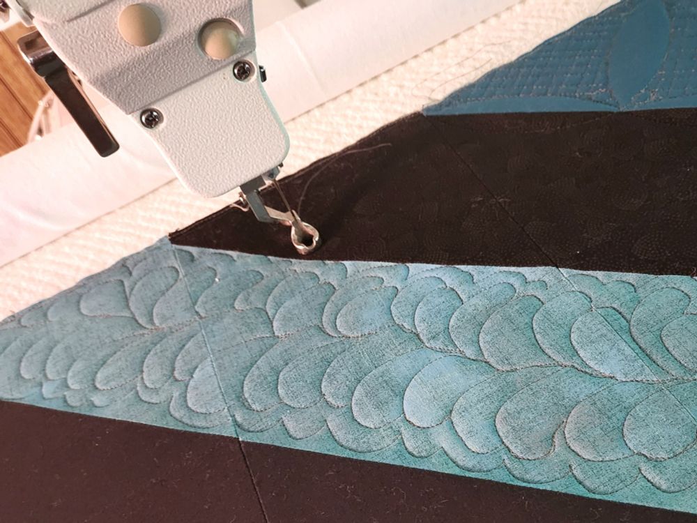 Photo of a teal blue and dark gray quilt on a longarm machine.  Shows the hand-guided quilting,  or stitching,  that creates an organic pattern 