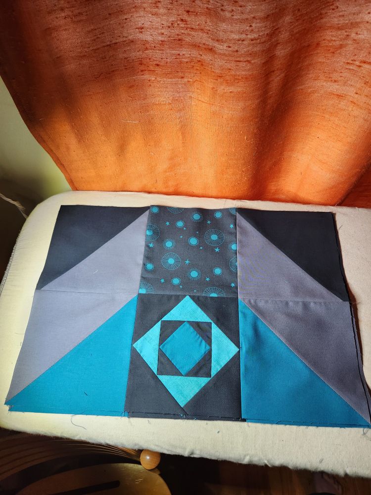 A teal blue and dark gray quilt block on an ironing board in front of a window with an orange curtain 