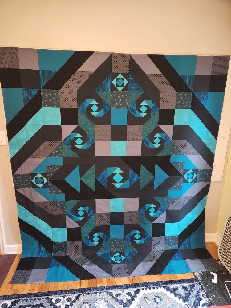 A quilt top made of dark gray and teal blue fabrics hanging on a design wall.  The bottom edge is laying on the floor. 