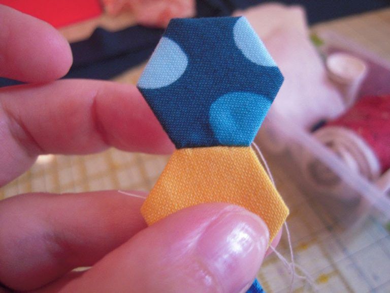 Close up of a hand holding two stitched hexie pieces from an English Paper Piecing project. The top hexie is blue with polka dots and the bottom hexie is yellow.