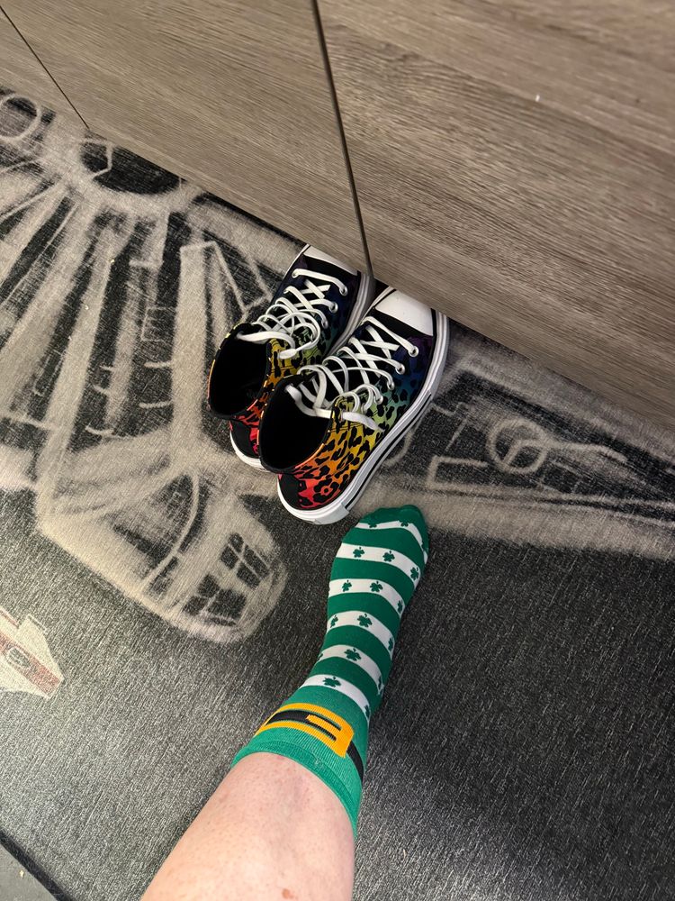 Rainbow converse shoes next to foot with a boot buckle and shamrock sock all which rest on a star wars rug