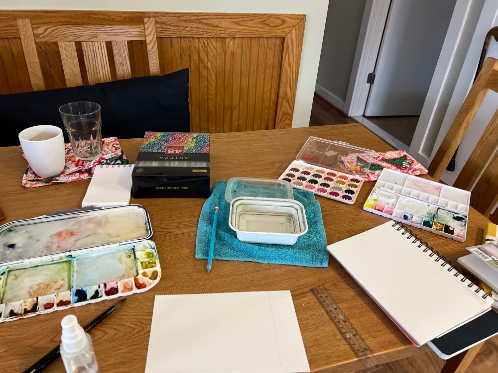 Table covered in various art supplies:sketchbooks, palettes, watercolor markers, ruler, brushes, water. 
