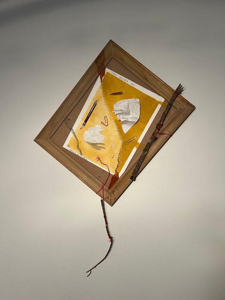 Crookedly hung art piece with real and painted office supplies and sticks. One dangling below the frame. 