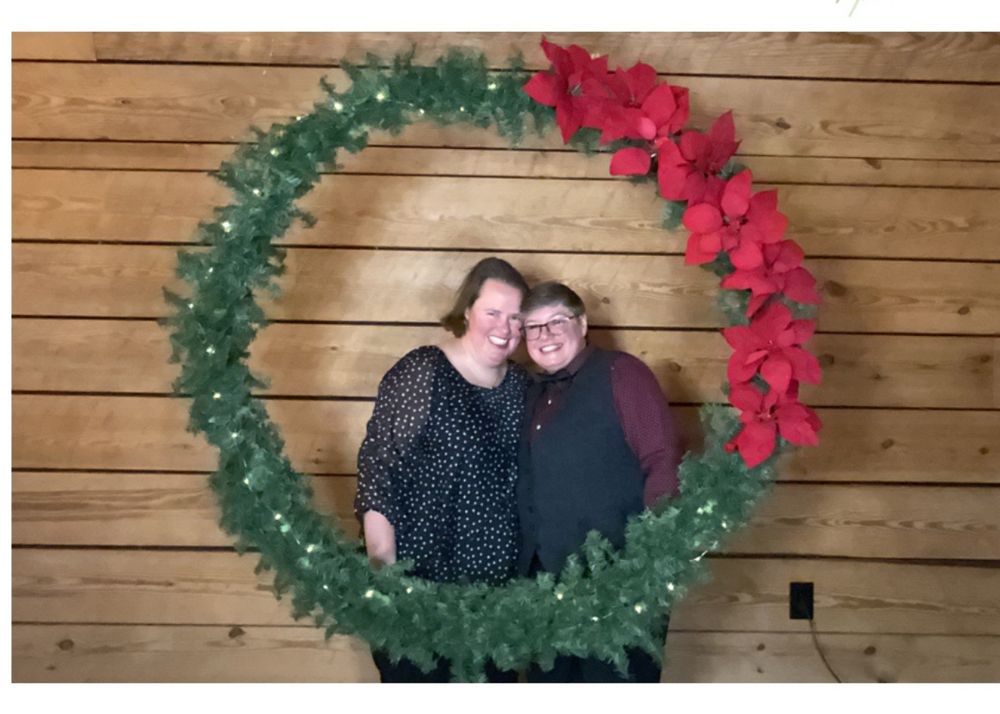 Two women in dark attire framed by a wreath with a wooden background. 