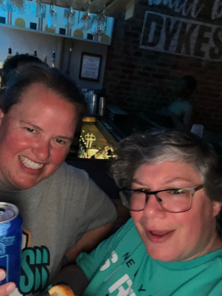 Two women toasting in front of a sign that says Dykes