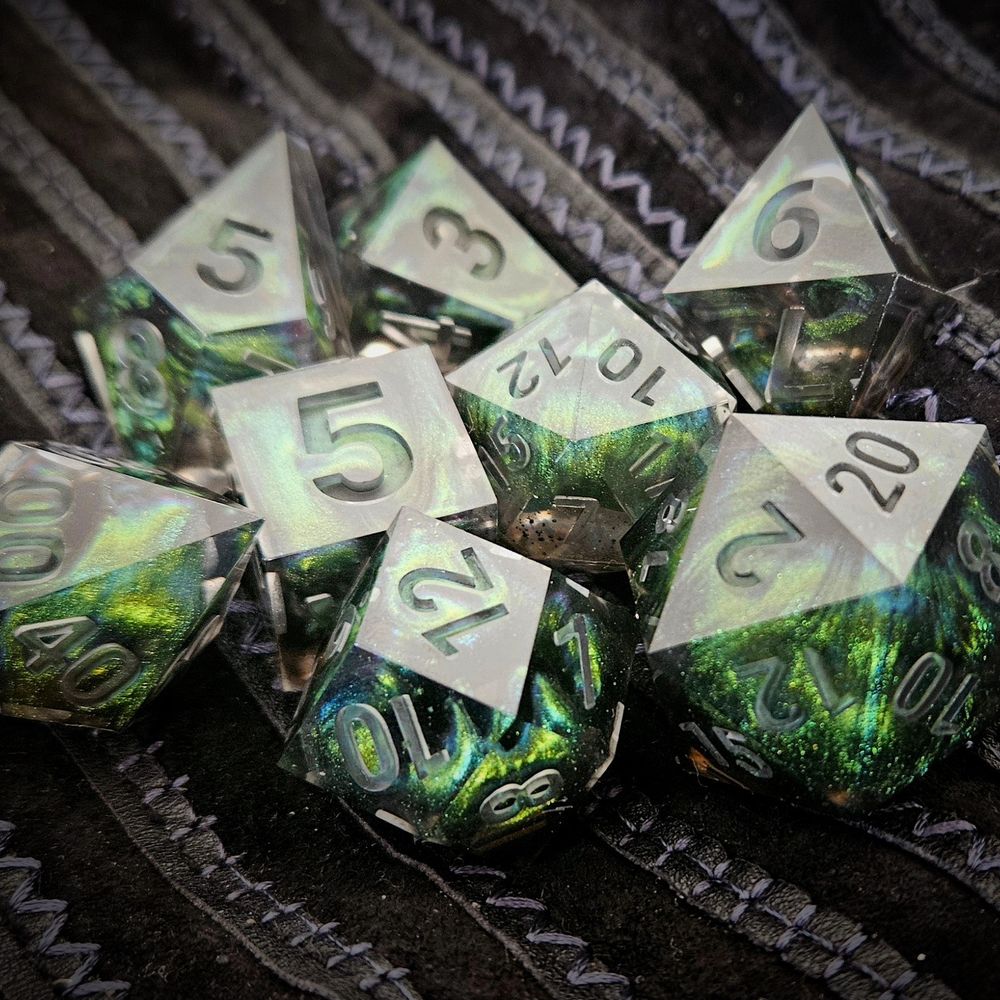 A high-angle photo of 8 handmade resin tabletop dice on a black suede background (a classic 7-piece tabletop RPG set and one 30mm D20). The dice are shiny and have a green/blue/purple colorshift effect. 