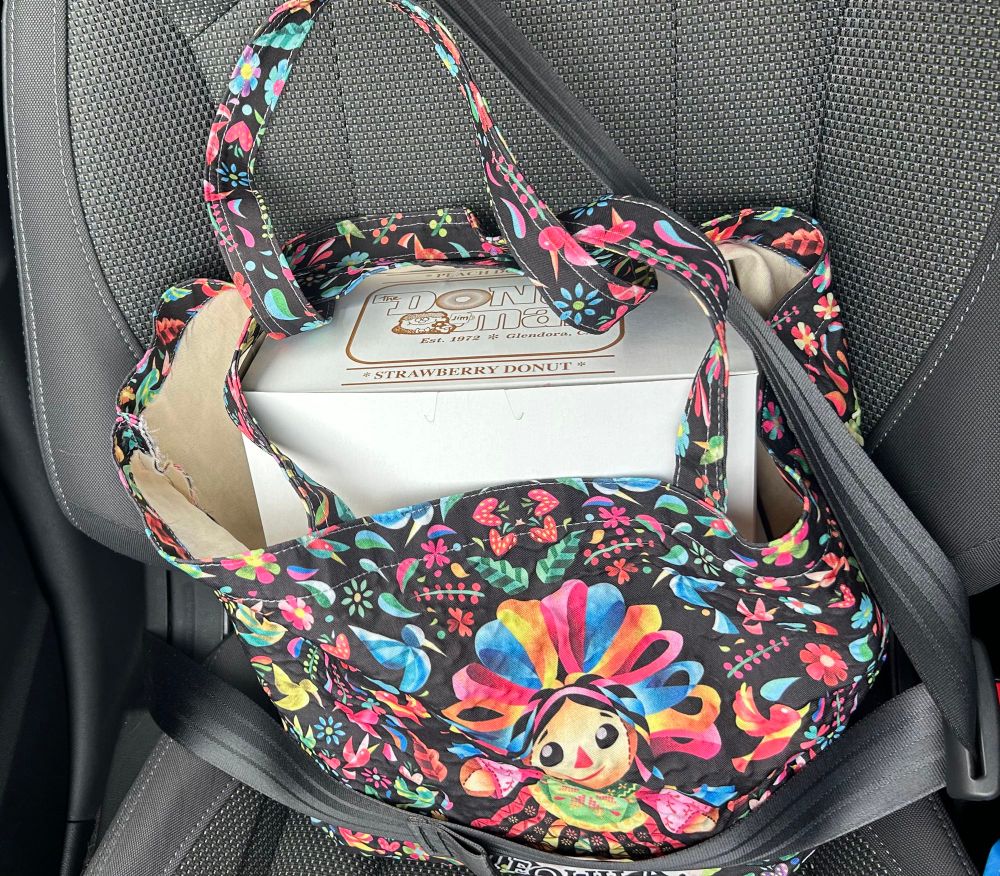 Reusable colorful bag with print of Mexican doll. Bag contains a Doughnut Man Box and is strapped on to the passenger side seat with a seatbelt.