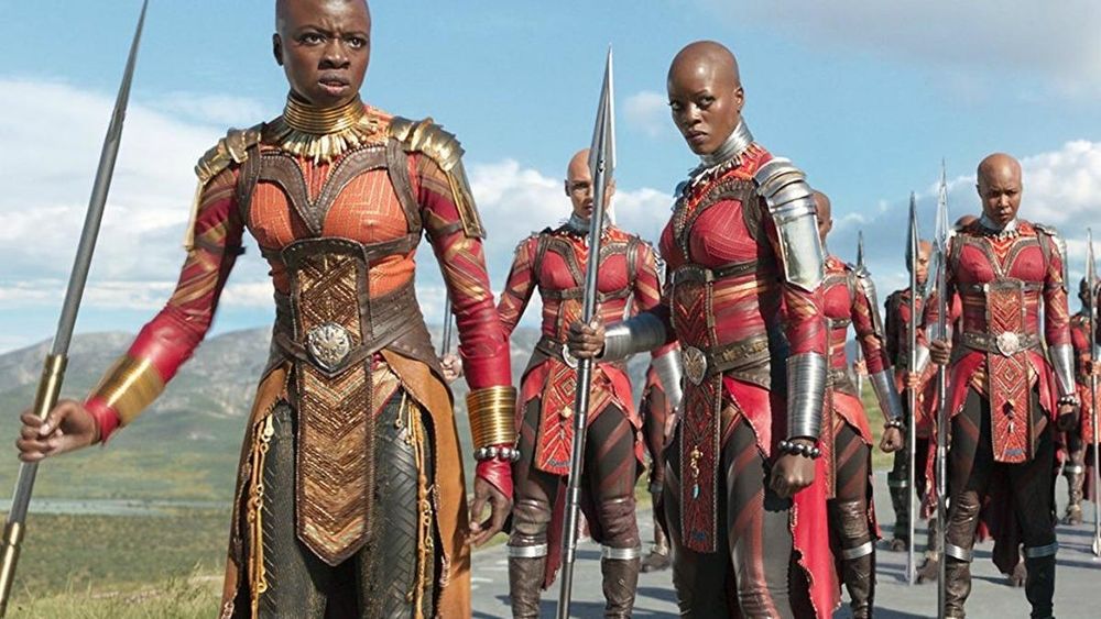 A still from the film Black Panther showing the Dora Milaje looking tough, holding their spears in their red leather armor against a blue sky.   