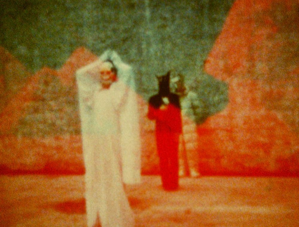 A grainy, blurry image of three figures. In the foreground, a figure dressed in a long white dress and cape and wearing a skull mask stands with their hands above their heads. Behind them, there is a figure wearing a long black dress and a headpiece with black horns. Behind them, just visible, is a third figure wearing what appears to be a padded suit and some kind of helmet. The background is orange and suggestive of pyramids and sand. The sky is a teal colour.

