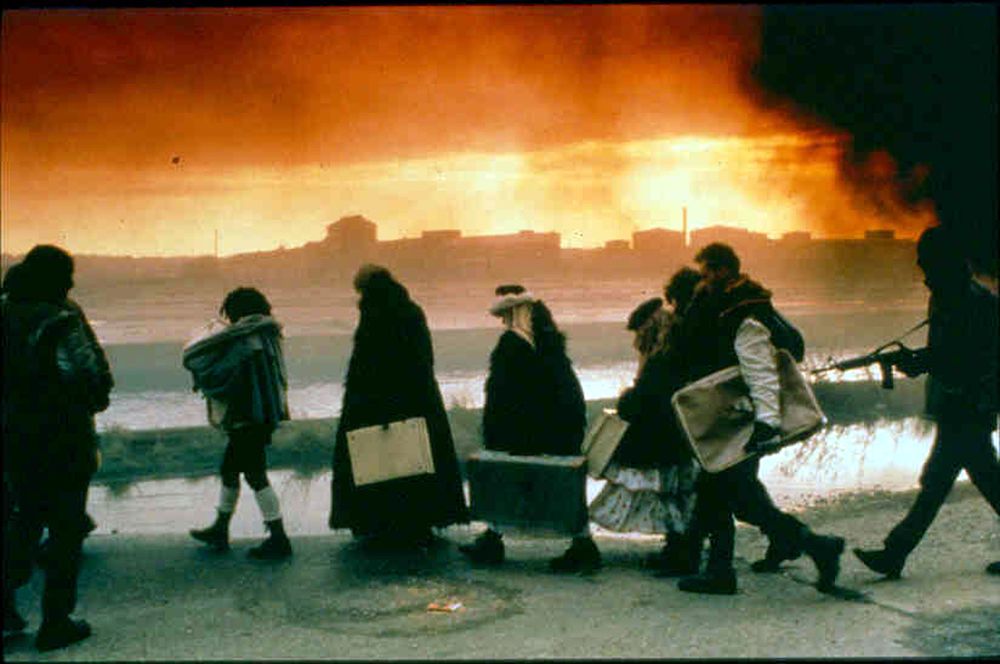 A group of people carrying luggage with soldiers pointing guns at them. They are silhouetted against a skyline with a yellow, orange and black sky above, suggesting fire.