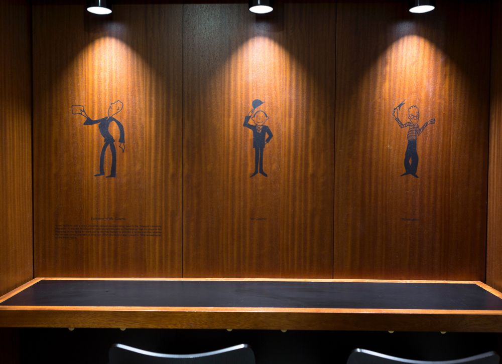 A dark wood panelled wall in GFT featuring three illustrations of a cartoonish male figure. In the first image, he is wearing a suit and brandishing a cinema ticket. In the second, he is wearing a suit and lifting a bowler hat. The third image shows a different man who has spiky hair, glasses and a striped top. Each image has a spotlight illuminating it from above.