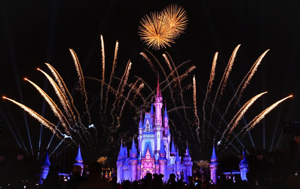A brightly illuminated castle, lit with blue and purple hues, serves as the centerpiece of the image, surrounded by a night sky. Fireworks burst above and around the castle in golden arcs, creating a stunning visual display. The silhouettes of a crowd in the foreground watch the spectacle, adding to the atmosphere of excitement and wonder. The setting is part of a celebratory or theme park event at Disney World.