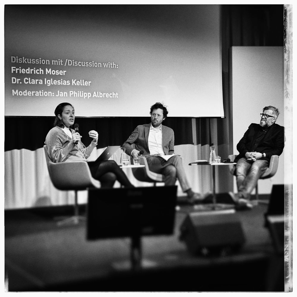 An indoor discussion panel featuring three speakers: a woman (Dr. Clara Iglesias Keller) speaking while holding notes, a man (Jan Philipp Albrecht) in a light suit listening, and another man (Friedrich Moser) in a dark jacket and glasses. A screen in the background displays the names of the panel participants. The image is in black and white.
