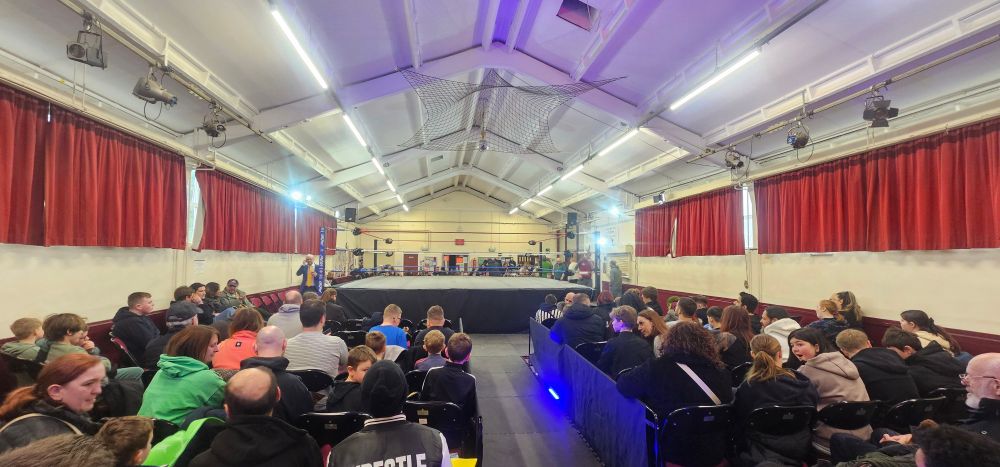 The setup for UKPW Blizzard Brawl 4. The photographer is seated in the back row of the section, and this wide shot shows a large crowd, reflecting how popular the wrestling is in Falconwood. The ring and wrestler entrance aisle are prominent in the centre of the picture. The wrestlers' entrance and commentary position are on the venue's stage, immediately behind the photographer. 