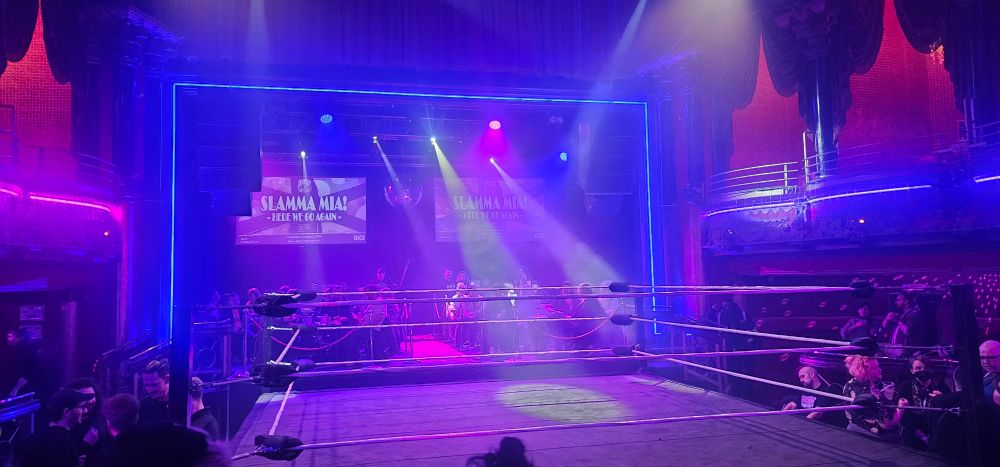 A shot of the Clapham Grand set up for a Riot Cabaret show. There are tables set up on the stage flanking the wrestlers entrance. The ring is surrounded by ringside seats, with the photographer in a raised standing area behind another row of  seats.