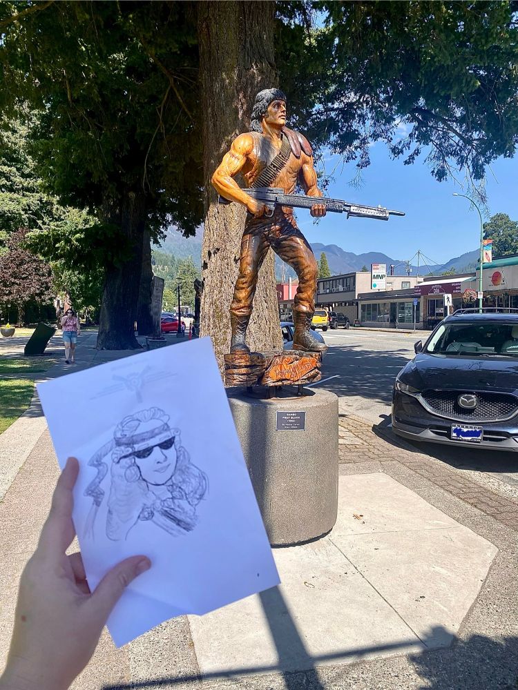 A crude hand drawing of the logo for worst of all possible worlds is held in front of a carved statue of Rambo