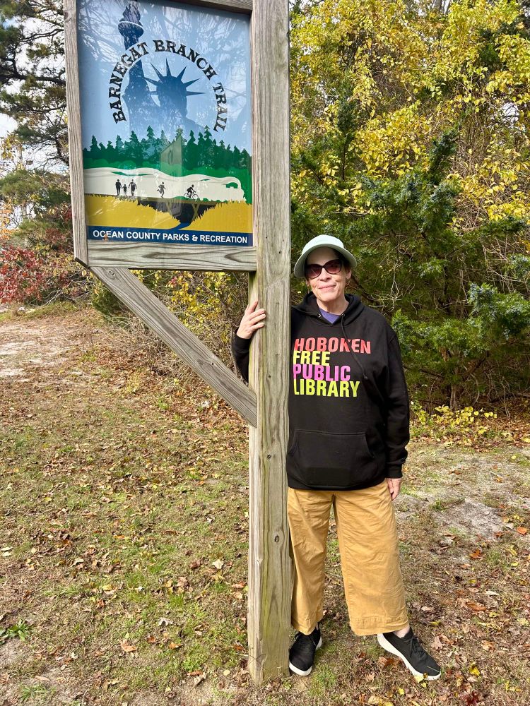 Woman wearing a Hoboken Free Public Library sweatshirt stands next to sign for the Barnegat Branch Trail