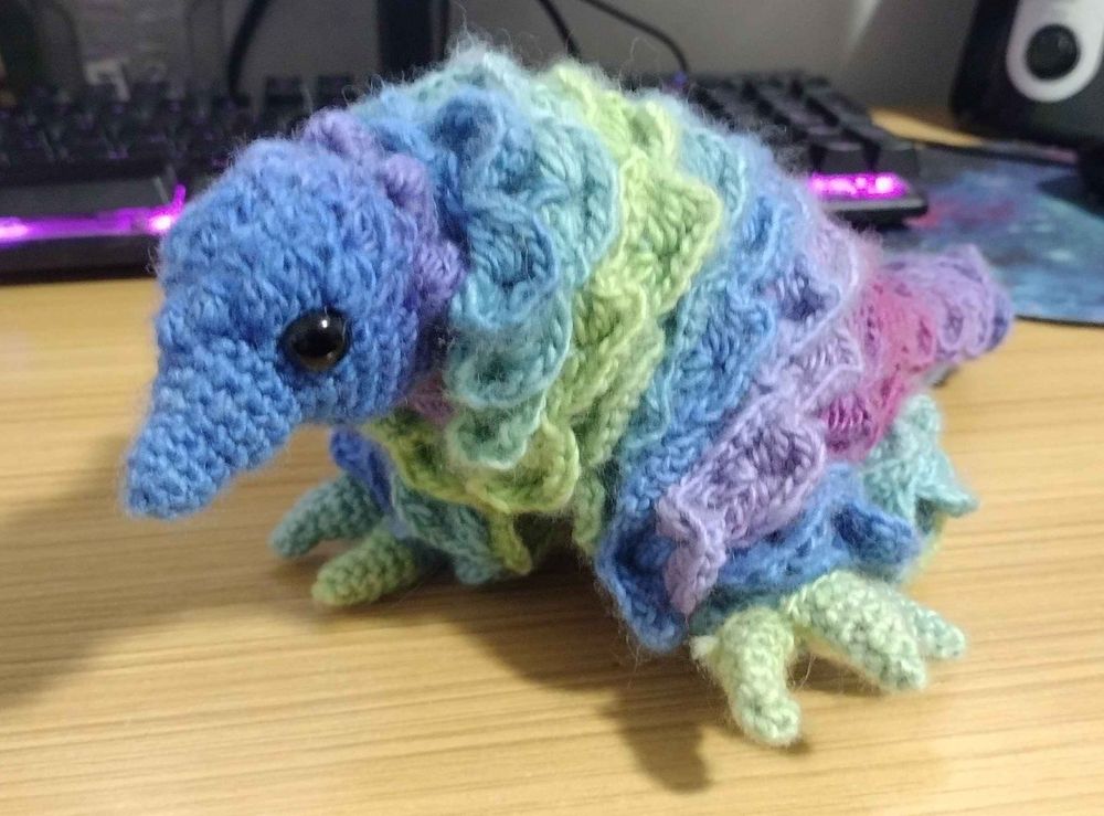 A photo of the crocheted pangolin, a pointy nosed creature with a scale-covered body, large digging hands. The yarn used is multicolored, ranging from violet to lavender to medium blue to aquamarine to lime and back through those colors. It sits on a background of a desk with a glowing keyboard.