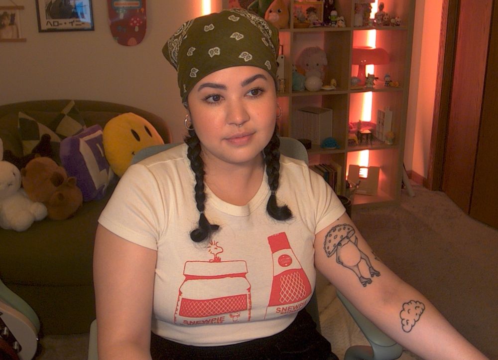 a screenshot of orzul, a biracial cambodian-american. their hair is black and styled in pigtail braids and wearing a green bandana. the background is filled with a chair, bookshelf, lots of stuffed animals, trinkets, and artwork. there is a soft warm huge from the lights. 

