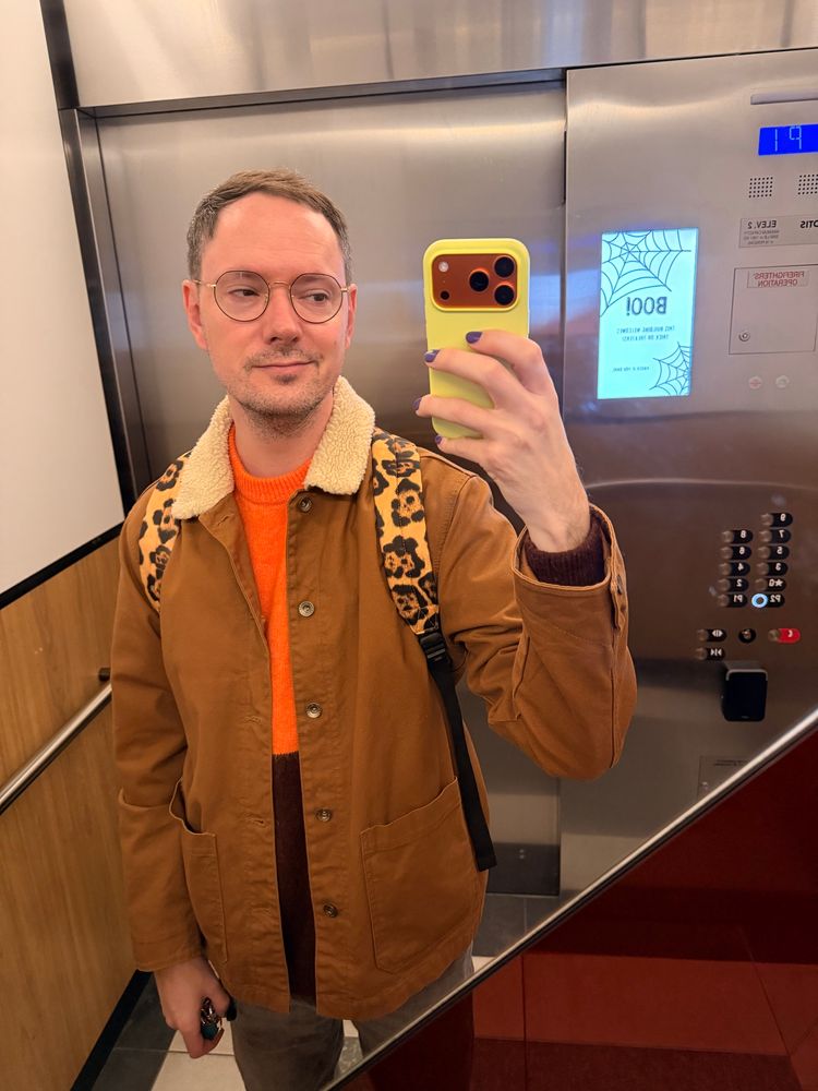 A man taking a selfie inside an elevator. He’s wearing a brown shearling coat, glasses, grey jeans and an orange/maroon sweater.