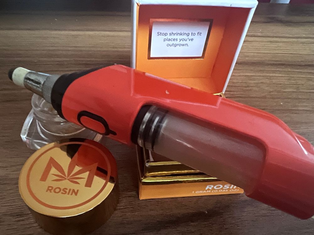An open orange, gold, and white Dime Industries Rosin box sits on a wooden table with the open jar and too next to it. Atop is the orange Looka Seahorse Pro dab rig.