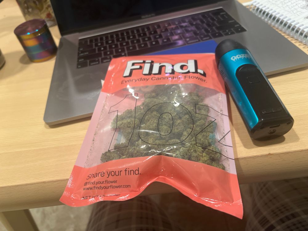 1oz bag of cannabis next to a blue Verge Dry Herb Vape laying atop a MacBook Pro. There is a rainbow metallic weed grinder to the left of the macbook pro.