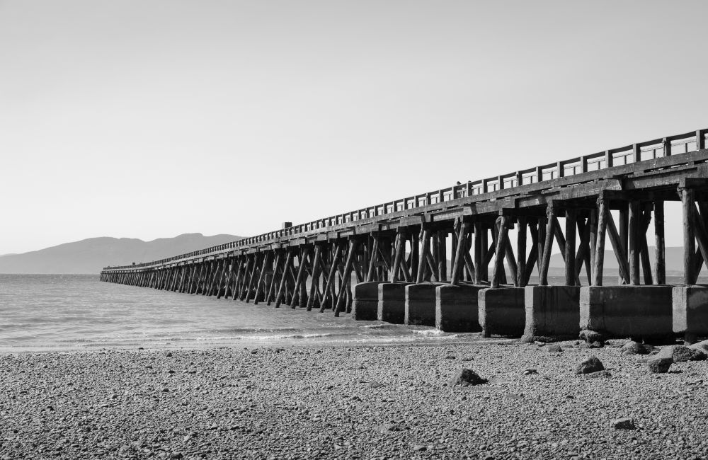 At the Bellingham waterfront, a long wooden pier stretches across the bay toward the hazy outline of Lummi Island. Pebbles and drift rocks fill the quiet shoreline beneath a pale, cloudless sky.