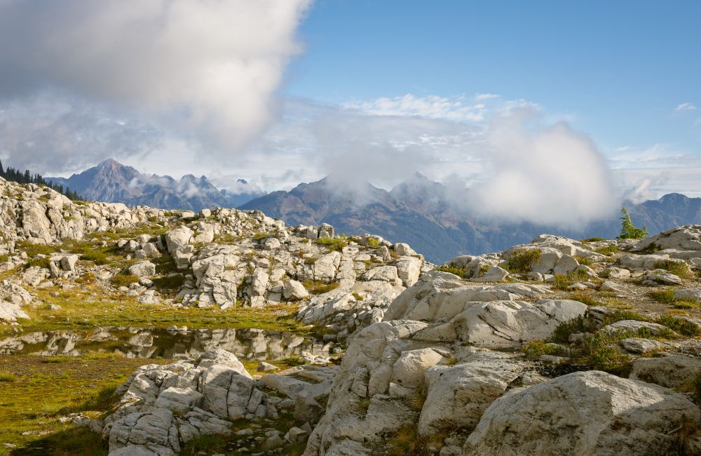Sunlit white granite slabs spread across a high ridge in the Mount Baker Wilderness. A shallow alpine pool reflects the rocks and patches of green meadow. Low clouds drift around jagged North Cascades peaks in the distance under a clear blue sky.