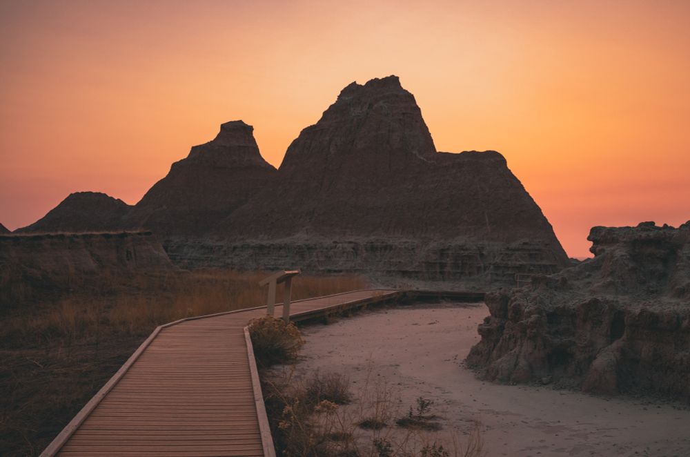 A wooden boardwalk winds through the dramatic terrain of Badlands National Park at sunrise. Jagged rock formations rise in silhouette against a vivid orange and pink sky, while dry grasses and rugged earth frame the quiet desert scene.