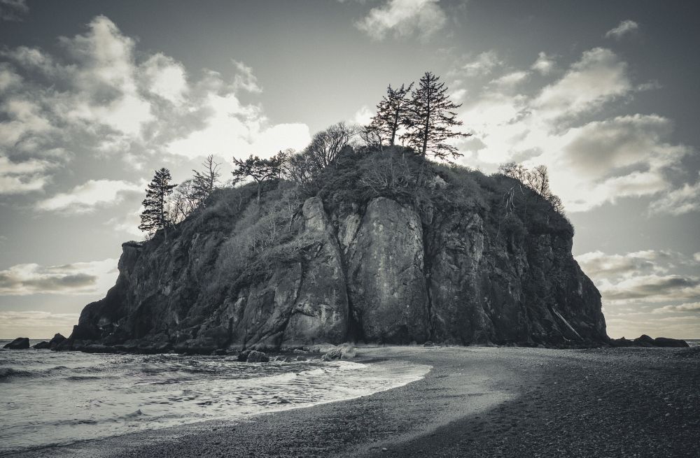 A rugged sea stack rises from the shoreline, its dark rock face streaked with age and topped with wind-shaped evergreens. Soft waves curl around the base under a sky streaked with pale clouds and fading afternoon light.