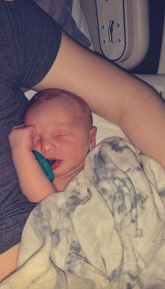 My daughter as a newborn baby cuddled up to my side with one arm up by her face, her soother is hanging out of her mouth a bit and she’s under a white and grey marbled blanket 