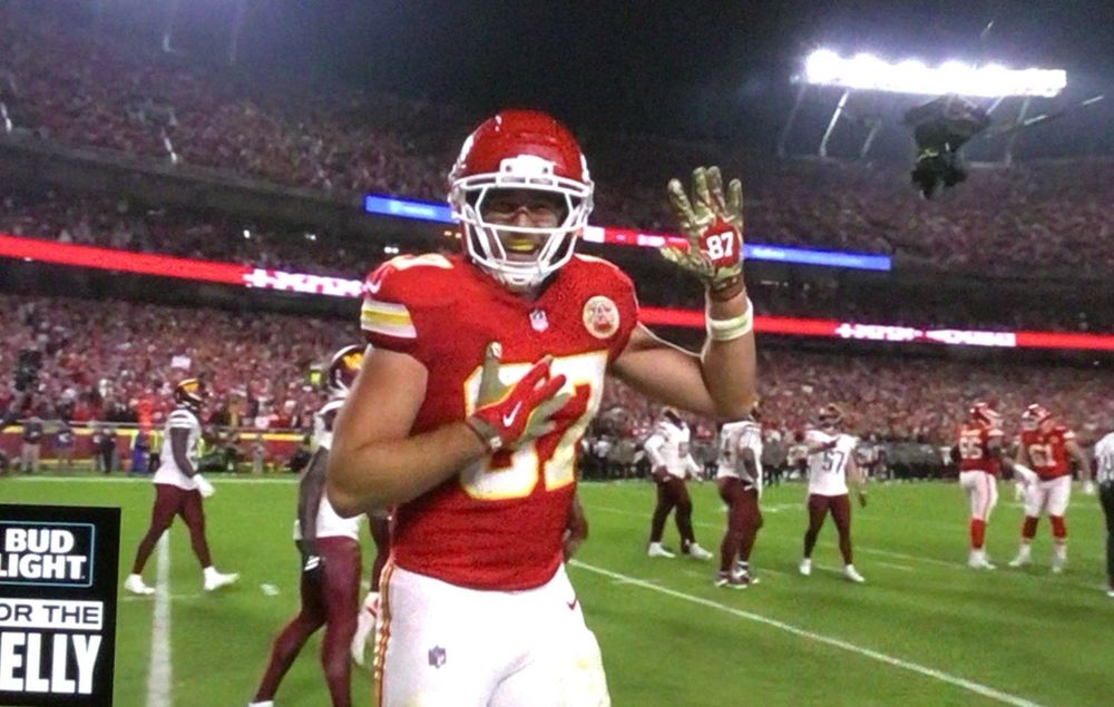 Travis Kelce with one hand on his heart and the other held up showing the 87 on his glove as he celebrates a touchdown 