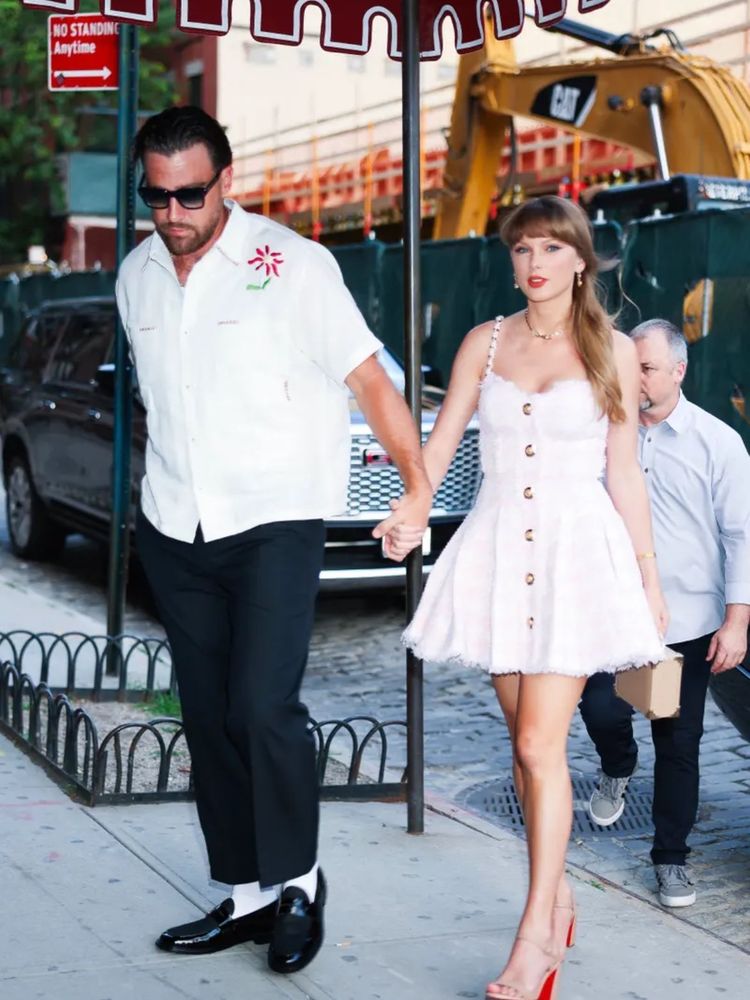Travis Kelce and Taylor Swift walking hand in hand. He’s wearing black dress pants and dress shoes and a white collard shirt with a red flower on the top left. He also has sunglasses on. Taylor is wearing a pink/white checkered dress with buttons down the front and red bottom heels. 