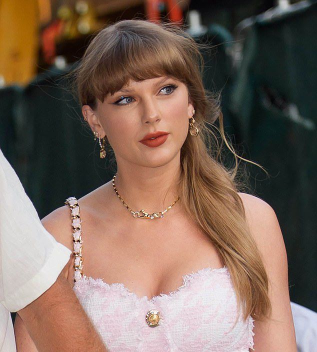 Taylor Swift wearing a pink/white checkered dress with a button at the top and gold accents on the straps. She had gold jewelry on it and red lipstick. 