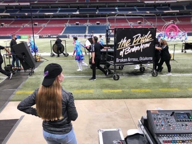 The account owner on the sideline of a football field watching a marching band set up. The drum major stand says “The Pride of Broken Arrow” and members in uniform can be seen moving into place. 